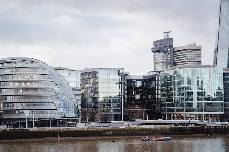 Exterior view of modern commercial office buildings with glass facades, situated along a riverbank. The buildings feature reflective windows that mirror the cloudy sky above, and some structures have